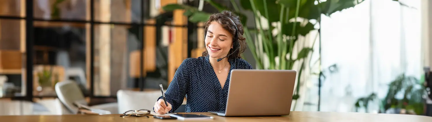 Eine Frau in gepunkteter Bluse sitzt mit einem Headset am Laptop und notiert etwas auf Papier