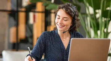 Eine Frau in gepunkteter Bluse sitzt mit einem Headset am Laptop und notiert etwas auf Papier
